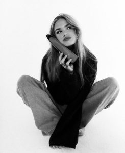 Young woman sitting cross-legged in casual denim and sweater, holding a ballerina flat to her ear like a phone, imitating to contact us, in a black-and-white editorial shot.